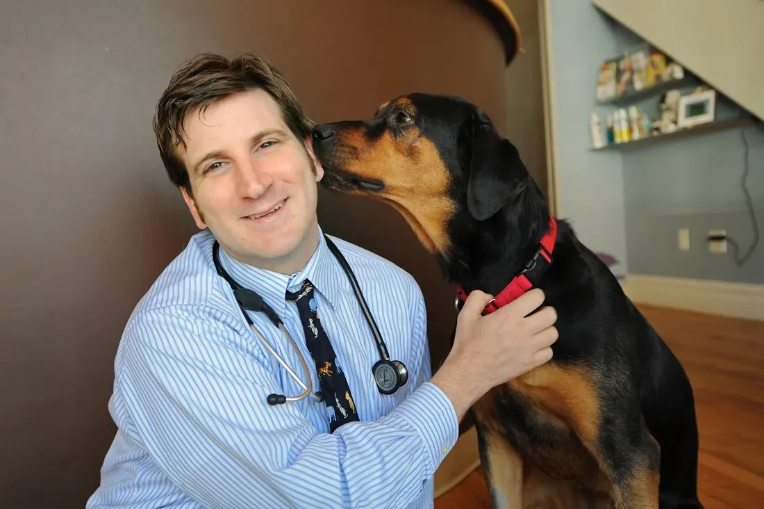 Dr. Timothy Mann in a striped shirt and stethoscope smiling as a black and tan dog in a red collar nuzzles his face